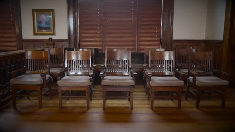 A photograph of the interior of a courtroom shows two rows of six empty chairs that make up a...